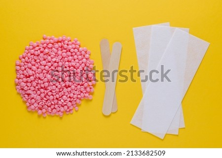 Beautiful pink depilatory wax granules, strips for depilation and wooden spatulas on a yellow background. Epilation, depilation, unwanted hair removal. Top view.