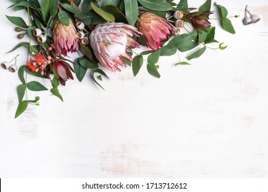 Beautiful Pink King Protea Surrounded By  Pink Ice Proteas, Leucadendrons, Eucalyptus Leaves And Flowering Gum Nuts, Creating A Floral Border On A Rustic White Background. Space For Copy.