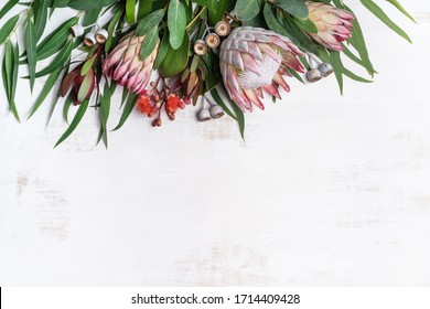 Beautiful Pink King Protea Surrounded By  Pink Ice Proteas, Leucadendrons, Eucalyptus Leaves And Flowering Gum Nuts, Creating A Floral Border On A Rustic White Background. Space For Copy.