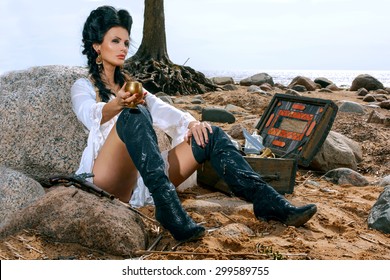 Beautiful pirate woman sitting near treasure chest on the beach with a golden goblet in her hand