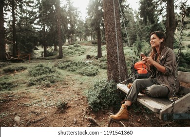 Beautiful place. Portrait of charming girl sitting on wooden swing and enjoying favorite song. She is holding mobile phone and looking away with smile - Shutterstock ID 1191541360