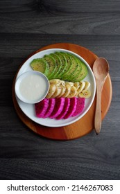 A beautiful plate of fruit salad, such as avocado slices, banana slices and dragon fruit slice. Sprinkle with chia seed, serve with greek yoghurt on a white plate and wooden chopping board. Isolated. 