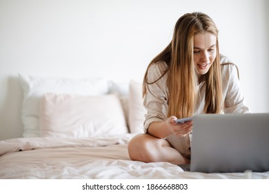 Beautiful pleased nice girl using laptop and cellphone in bed at home