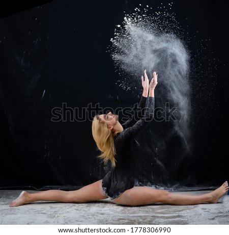 Beautiful plump blonde girl wearing a black gymnastic bodysuit covered with clouds of the flying white powder  dancing sitting on a dark. Artistic conceptual and advertising photo. Copy space