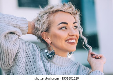 Beautiful Plus Size Model With Blond Curly Hair Wearing Grey Knitted Sweater And Silver Earrings Posing On City Street. Fashion Everyday Outfit For Cold Season.