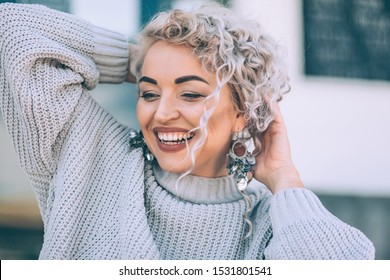 Beautiful Plus Size Model With Blond Curly Hair Wearing Grey Knitted Sweater And Silver Earrings Posing On City Street. Fashion Everyday Outfit For Cold Season.