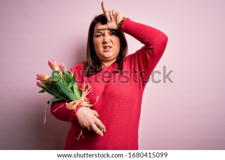 Beautiful plus size woman holding romantic bouquet of natural tulips flowers over pink background making fun of people with fingers on forehead doing loser gesture mocking and insulting.