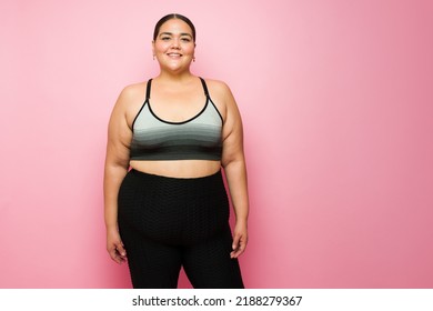 Beautiful plus size woman smiling wearing activewear and ready for exercise against a pink studio background