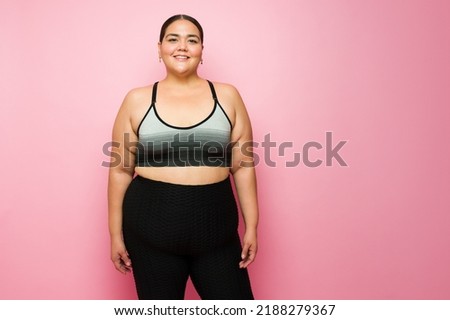 Beautiful plus size woman smiling wearing activewear and ready for exercise against a pink studio background