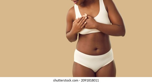 Beautiful Plus Size Young Black Woman In Underwear Touches Her Chest When She Feels Strong Pain Standing Isolated On Beige Color Banner Background. Breast Cancer And Heart Attack Awareness Concept