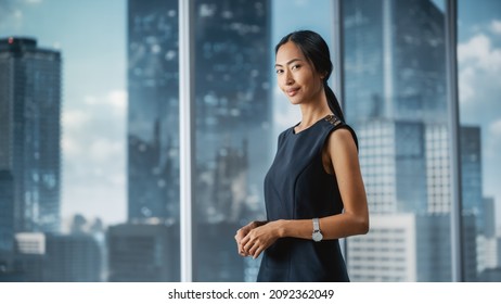 Beautiful Portrait Of An Asian Businesswoman In Stylish Black Dress Posing Next To Window In Big City Office With Skyscrapers. Confident Female CEO Smiling. Successful Diverse Business Manager.