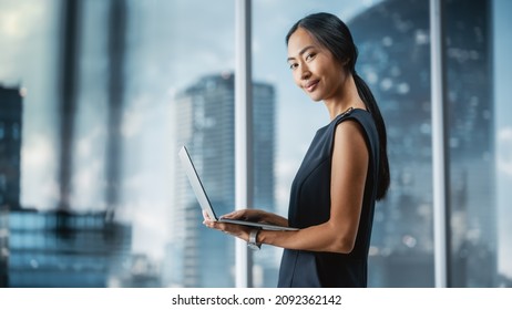 Beautiful Portrait Of An Asian Businesswoman In Stylish Black Dress Using Laptop Computer, Posing Next To Window In City Office. Confident Female CEO Smiling. Successful Diverse Business Manager.