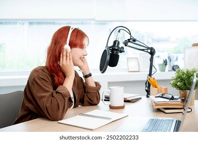 A beautiful and positive Asian female podcaster with red curly hair, wearing headphones, is recording her podcast in the studio, speaking into the microphone, creating an engaging audio podcast. - Powered by Shutterstock