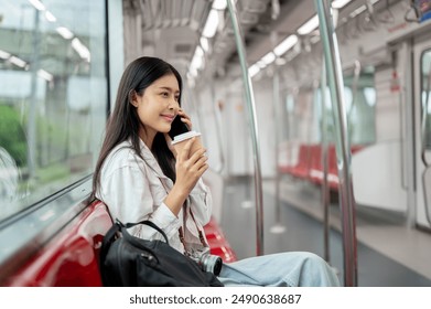 A beautiful, positive Asian female tourist with a backpack is sipping coffee and talking on the phone while commuting on a sky train. people and public transport concepts - Powered by Shutterstock