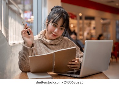 A beautiful and positive Asian woman in a cosy sweater is working remotely at a coffee shop, multitasking, listing her ideas in a book and working on her laptop. - Powered by Shutterstock