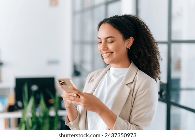 A beautiful positive brazilian or hispanic young woman with curly hair in an elegant jacket, stands in a modern office space, uses her smartphone, messaging with friends on social networks, smiles - Powered by Shutterstock