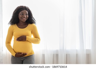 Beautiful pregnant african american woman hugging her big belly, enjoying her pregnancy, looking at free space. Young black expecting lady standing next to window at home, touching her tummy
