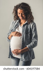 Beautiful pregnant Afro American woman is touching her tummy and smiling, on gray background