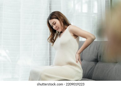 Beautiful Pregnant Asian Woman Is Sitting On A Couch In The Living Room, Having A Back Pain As She Carries The Baby In Her Tummy. Pregnant Problem.