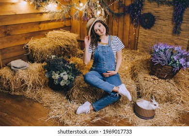 A beautiful pregnant brunette girl in a straw hat and denim overalls is sitting posing in the hay with decorations, a rabbit, flowers and a gerland.