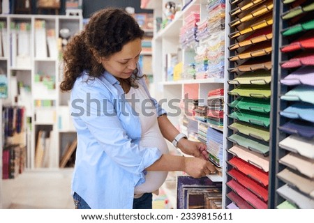 Beautiful pregnant woman, customer choosing vibrant color paper for design, fine art or painting, standing by shelves with cardboard sheets in creative art shop of stationary store