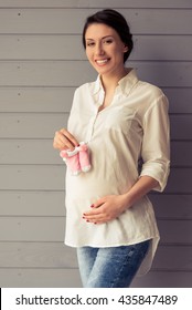 Beautiful Pregnant Woman Is Holding Baby Shoes, Keeping One Hand On A Belly, Looking At Camera And Smiling, Standing Against Gray Wall