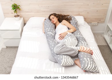Beautiful pregnant woman with long curly hair and a big tummy sleeping on the bed by the window indoor and hugging his hand and knees convenient  ergonomic anatomic body-pillow.