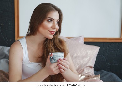Beautiful pregnant woman with long hair in a white T-shirt and pink pajamas sitting on the bed, holding a cup of tea. Concept of happy motherhood, healthy lifestyle.