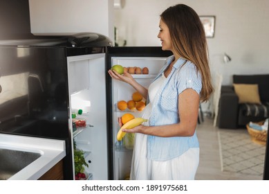 Beautiful pregnant woman looking into fridge, choosing fruits and vegetables for making vitamin smoothie. Healthy lifestyle in pregnancy.