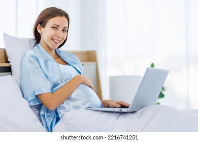 Beautiful Pregnant Woman  Pajamas Resting On Bed With Smile And Happy To Watch Laptop For Prenatal Care Of Maternity. Healthy Mom Relax And Pleased With Belly Of Unborn Baby For Expecting Child Birth.