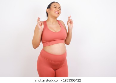 Beautiful Pregnant Woman In Sports Clothes Against White Background Has Big Hope, Crosses Fingers, Believes In Good Fortune, Smiles Broadly. People And Wish Concept