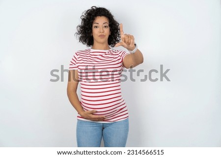 Beautiful pregnant woman standing over white studio background making fun of people with fingers on forehead doing loser gesture mocking and insulting.