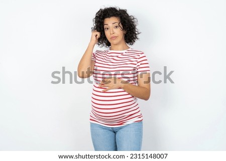 Beautiful pregnant woman standing over white studio background tries to memorize something, keeps fore finger on temple, reminds information for exam,