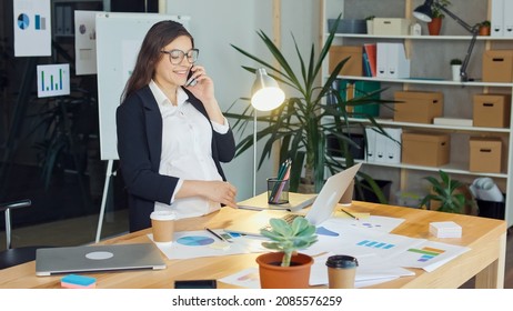 A Beautiful Pregnant Woman Talking on a Cell Phone Sitting at a Table in a Comfortable Modern Office, Gently Stroking Her Tummy. Happy Modern Working Woman and Motherhood.
