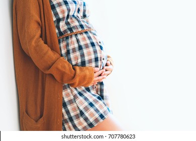 Beautiful pregnant woman touching her belly with hands on a white background. Young mother anticipation of the baby. Image of pregnancy and maternity. Close-up, copy space, indoors. Maternity wear