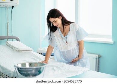 Beautiful Pregnant Woman Trying To Breath Deeply For Relieve Pain Of Labour Contractions At Hospital. Young Woman Standing At Medical Ward And Leaning With Hands On Couch.