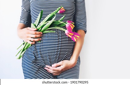Beautiful pregnant woman with tulips flowers holds hands on belly on a white background. Stylish fashion woman in maternity dress waiting for a baby birth. Pregnancy, Mother's Day Holiday concept.