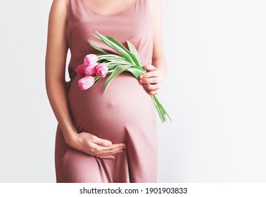 Beautiful pregnant woman with tulips flowers holds hands on belly in white background. Young woman in maternity pink dress waiting for baby birth. Pregnancy, Motherhood, Mother's Day Holiday concept.