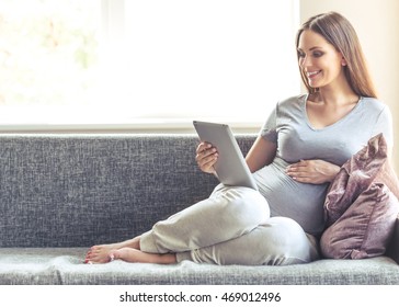 Beautiful Pregnant Woman Is Using A Digital Tablet, Keeping One Hand On Her Tummy And Smiling While Sitting On The Couch At Home