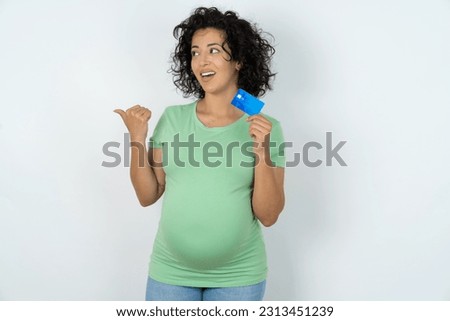 Beautiful pregnant woman wearing green T-shirt standing over white studio background hold credit card point empty space 