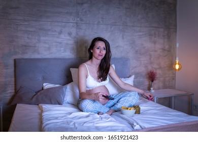 Beautiful pregnant woman wearing pajamas sitting on bed at night, switching channels using remote control and watching TV - Shutterstock ID 1651921423