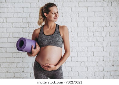 Beautiful Pregnant Woman Workout. Doing Fitness On Last Months Of Pregnancy. Yoga. Standing On White Brick Background With Sports Mat.