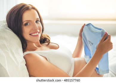 Beautiful Pregnant Young Woman Is Holding An Onesie, Looking At Camera And Smiling While Lying In Bed At Home