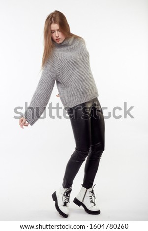 Beautiful   pretty girl with long brown hair and perfect body weared in black leather pants and gray sweater, down jacket, white boots. Isolated on white background.