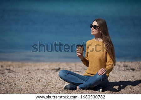 A beautiful pretty girl is sitting on the beach by the sea and drinking coffee to take away . The concept of natural beauty , relaxation, recreation, travel, coffee to take away . Copy space