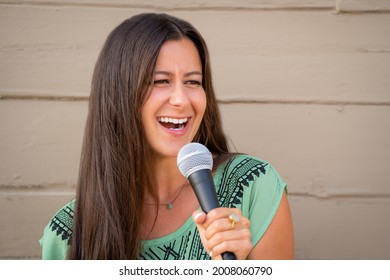 Beautiful Pretty Young Brunette Woman Stand Up Comedian With Microphone Telling Joke Outside On Street Against Wall Outdoors