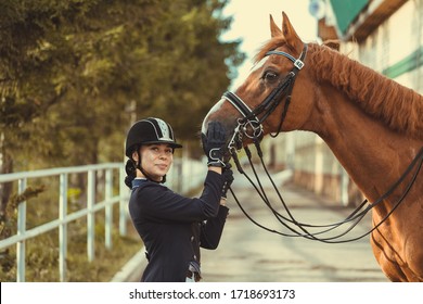 Beautiful Professional Female Jockey Standing Near Horse. Woman Horse Rider Is Preparing To Equitation. Girl And Horse. Equestrian Sport Concept. Dressage Horse 