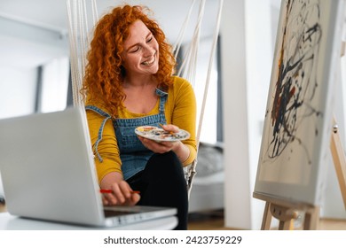 Beautiful quirky female artist, sitting alone in her art studio and smiling while painting and typing on laptop. Young woman multitasking at home, sitting on a rope swing. - Powered by Shutterstock