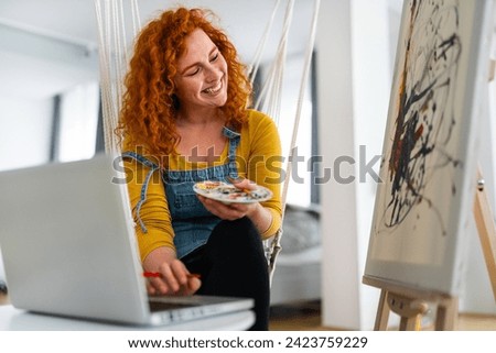Beautiful quirky female artist, sitting alone in her art studio and smiling while painting and typing on laptop. Young woman multitasking at home, sitting on a rope swing.