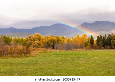 Beautiful Rainbow Rising Up From Colorful Fall Foliage With Mountains In Background And Farmer's Field In Foreground Near Whitefish, Montana